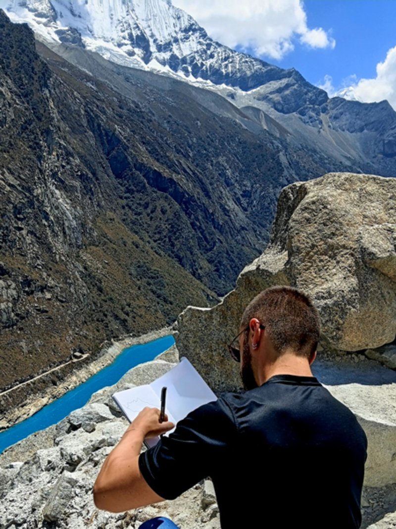 Felix Benito sketching in a notebook overlooking a glacial lake and snow-capped mountain in Peru
